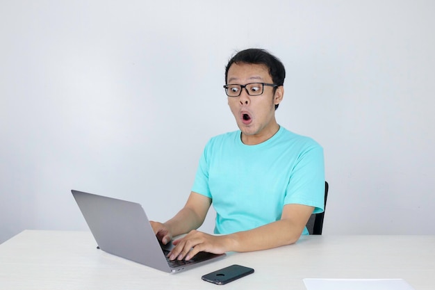 Wow, il volto del giovane asiatico ha scioccato ciò che vede nel laptop quando lavora su uno sfondo grigio isolato indossando una camicia blu