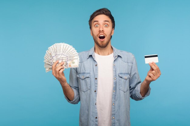 Wow, incredibile servizio di trasferimento di denaro. Uomo stupito in camicia di jeans da lavoratore che mostra banconote in dollari e carta di credito, guardando la fotocamera con la faccia scioccata. tiro in studio indoor isolato su sfondo blu