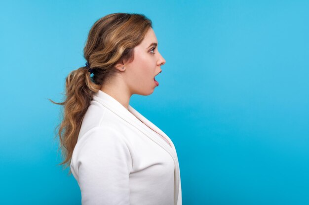 Wow, incredibile, vista laterale di una donna stupita con una pettinatura a coda di cavallo in giacca bianca che guarda con la bocca aperta per la sorpresa, scioccata da notizie pazze. studio indoor girato isolato su sfondo blu, copia spazio