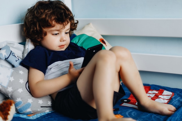 Wow, mi piace quel telefono. Bambino con smartphone. Ragazzo che si siede nel letto e che gioca con il telefono cellulare. Chiamando mia mamma. Carino piccolo bambino tiene il cellulare in mano e guardando attentamente lo schermo.