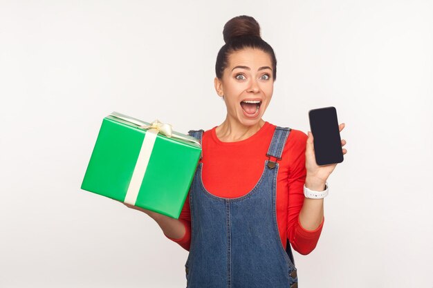 Wow, regalo per utenti mobili. Ritratto di una ragazza graziosa e gioiosa stupita con un panino per capelli in tuta di jeans con scatola regalo e telefono cellulare, scioccata dal piano tariffario e dal bonus delle vacanze. girato in studio, isolato