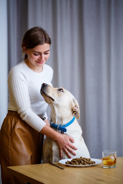 Yellow Labrador Retriever è seduto al tavolo con la sua amante. Cibo per cani in un piatto bianco, amico del cane dell'uomo