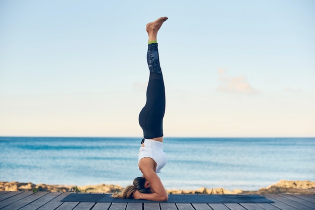 Yoga di pratica della bella donna di misura contro il mare blu che sta nella posa del headstand.