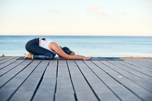 Yoga di pratica della bella giovane donna sulla spiaggia contro il mare blu.