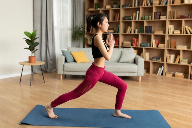 Yoga domestico Vista laterale della giovane signora giapponese che fa affondo tenendosi per mano in namaste che si esercita a casa a tutta lunghezza