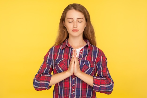 Yoga, pace interiore e armonia. Ritratto di felice ragazza zenzero calma in camicia a scacchi che prega con gli occhi chiusi, meditando tenendosi per mano nel gesto di namaste. girato in studio isolato su sfondo giallo