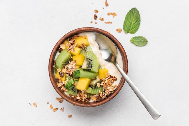 Yogurt al mango con muesli e kiwi in una ciotola di legno su sfondo bianco.