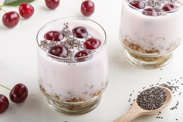 Yogurt con i semi e il granola di chia delle ciliege in vetro con il cucchiaio di legno sulla vista laterale del fondo di legno bianco