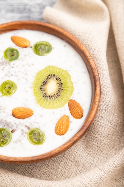 Yogurt con kiwi, uva spina, chia e mandorle in ciotola di legno su fondo di legno grigio e tessuto di lino. Vista dall'alto, primo piano, messa a fuoco selettiva.