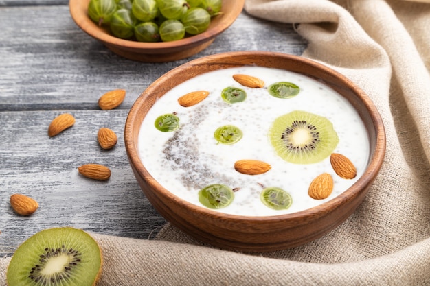 Yogurt con kiwi, uva spina, chia e mandorle in una ciotola di legno su fondo di legno grigio e tessuto di lino. Vista laterale, da vicino.