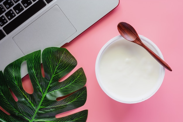 Yogurt con sapore naturale, cucchiaio di legno, foglia di palma verde e computer portatile sul bac rosa