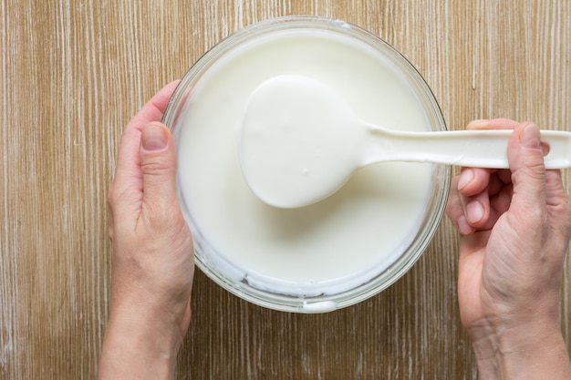 Yogurt greco in cucchiaio da cucina in mano di donna lo prese da una ciotola di vetro su sfondo di legno