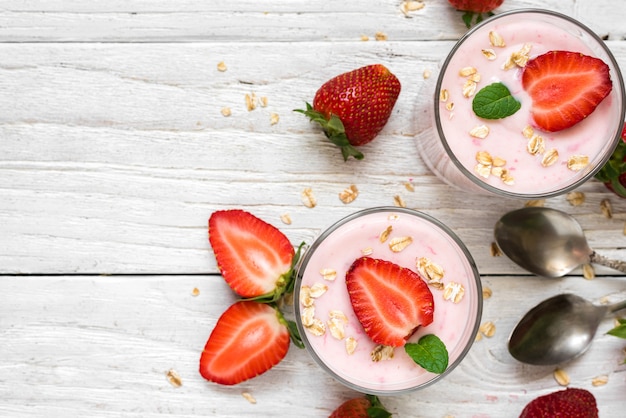 Yogurt sano della fragola con l'avena e la menta in vetri con le bacche e i cucchiai freschi sopra la tavola di legno bianca