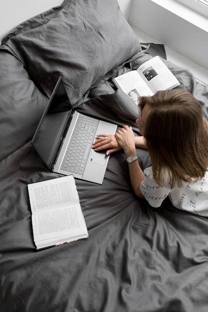 Yong donna sta lavorando o studiando sul letto con coperta grigio scuro portatile quaderno libro vista superiore