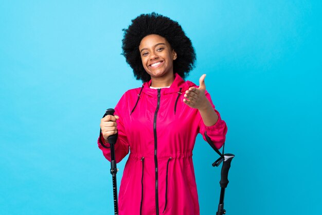 Young African American con zaino e bastoncini da trekking isolati su blu stringere la mano per chiudere molto