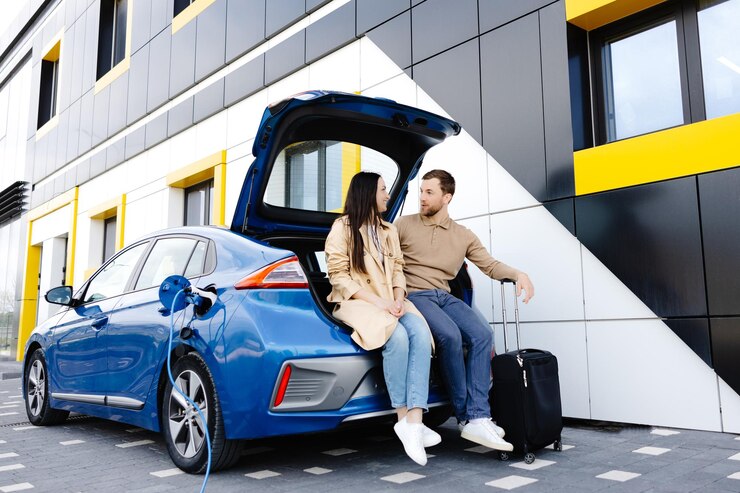 Young couple man and woman traveling by electric car having stop at charging station Boyfriend and girlfriend sitting in the car trunk and smiling