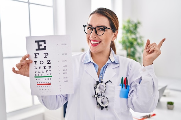 Young hispanic optician woman holding medical exam smiling happy pointing with hand and finger to the side