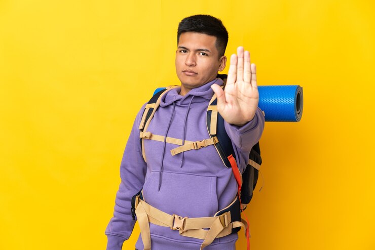 Photo young mountaineer man with a big backpack isolated on yellow wall making stop gesture