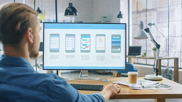 Young Professional Works on a Personal Computer at His Desk in the Creative Office Notebook Screen Shows Mobile Phone Application Design Software UI Development In Background