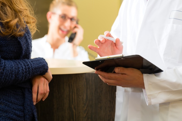 Zona paziente e dottoressa dell'ufficio del medico o del dentista, tiene in mano un blocco per appunti, l'addetto alla reception è al telefono