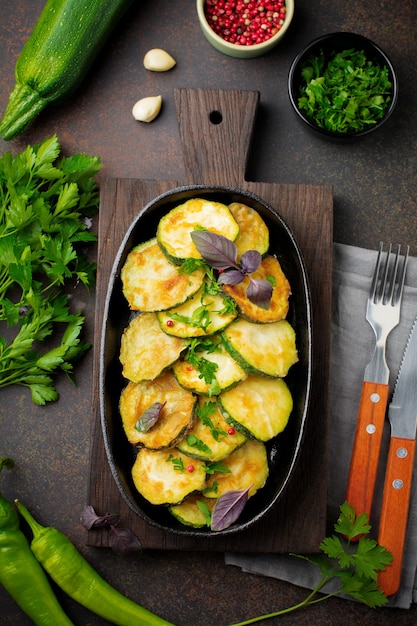 Zucchine fritte con peperoncino rosso, basilico e prezzemolo in una padella in ghisa sul vecchio sfondo scuro.