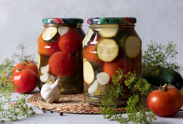 Zucchine marinate con pomodori in due vasetti di vetro per l'inverno su fondo grigio chiaro con ingredienti freschi