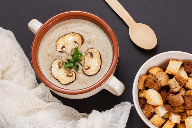 Zuppa di crema di funghi fatta in casa con funghi affettati in ciotola di porcellana, cucchiaio di legno e toast su sfondo nero. Vista dall'alto.