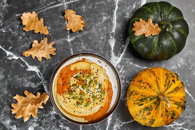 Zuppa di crema di zucca con panna e semi di zucca e cumino nero e zucche decorative organiche con foglie di quercia cadute.. Natura morta autunnale. Vista dall'alto