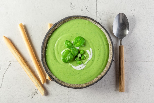 Zuppa di piselli e broccoli con crema verde sana con grissini italiani croccanti al basilico e grissini in una ciotola con cucchiaio su superficie di pietra chiara