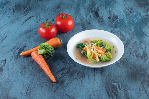 Zuppa di pollo con broccoli e carote in una ciotola accanto alle verdure, sulla superficie blu.