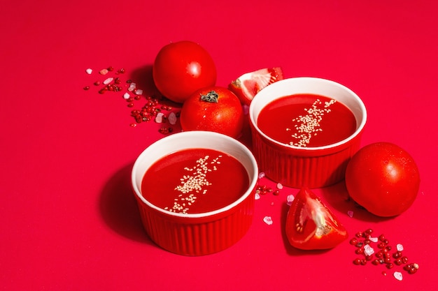 Zuppa di pomodoro con semi di sesamo in ciotole su fondo rosso. Verdure mature, sale rosa dell'Himalaya, pepe rosa in grani. Una moderna luce dura, ombra scura, copia spazio