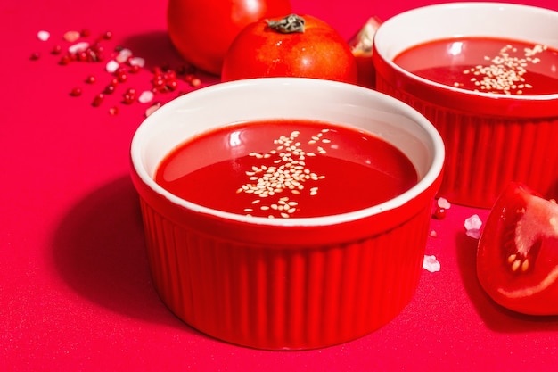 Zuppa di pomodoro con semi di sesamo in ciotole su fondo rosso. Verdure mature, sale rosa dell'Himalaya, pepe rosa in grani. Una moderna luce dura, ombra scura, primo piano