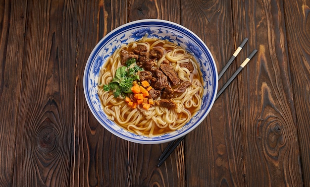 Zuppa di spaghetti di manzo cinese nella tradizionale ciotola blu, erbe aromatiche e carote affettate, coppia di bacchette sul tavolo in legno scuro e copia spazio. Vista dall'alto del concetto di cibo asiatico