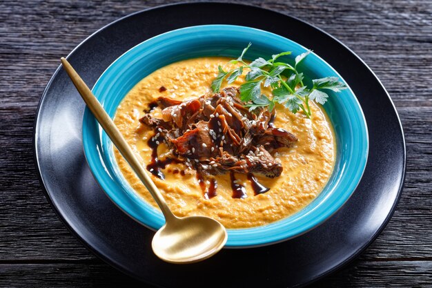 Zuppa di zucca di purea di zucca arrosto e brodo di funghi selvatici, condita con semi di sesamo, piatto vegano comfort, servito su un piatto blu con cucchiaio e aglio su uno sfondo di legno scuro, vista dall'alto