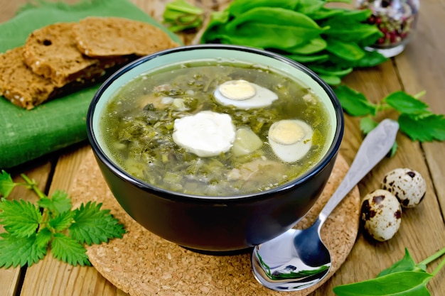 Zuppa verde di acetosa, ortiche e spinaci in una ciotola di uova di quaglia, pane, pepe, cucchiaio su una tavola di legno