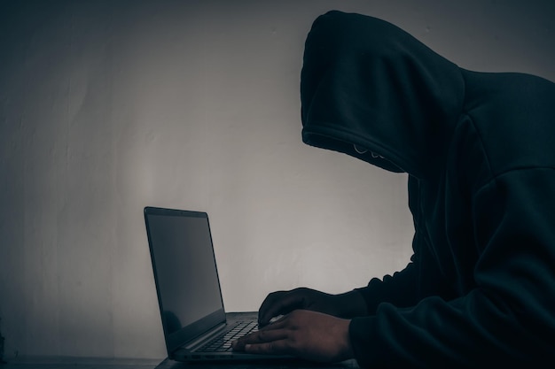 A hacker with a hood covering his head Sitting in a dark room using a laptop