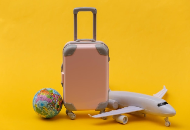 air travel concept. mini pink plastic travel suitcase, air plane, globe on yellow background.