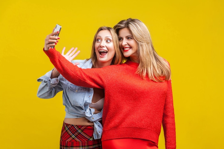 Fotografie best friends having fun. portrait of happy women in stylish clothes standing with toothy smile and taking selfie on phone together, enjoying life. indoor studio shot isolated on yellow background