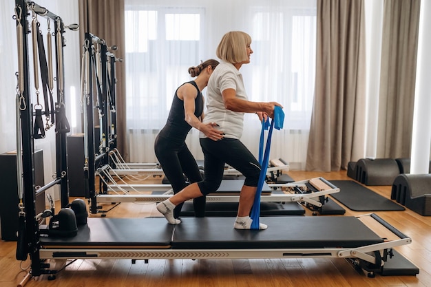 blue colored resistance loops in hands for the legs female trainer is helping old woman to exercise