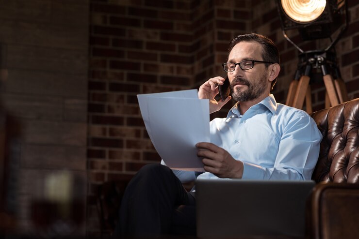 Fotografie call to a business partner. pleasant handsome delighted man putting a smartphone to his ear and talking to his business partner while holding some documents
