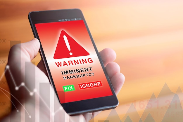 closeup of male hand holding modern mobile phone with red screen showing warning symbol and word with the text imminent bankruptcy accompanied by financial graphics concept of economic problems