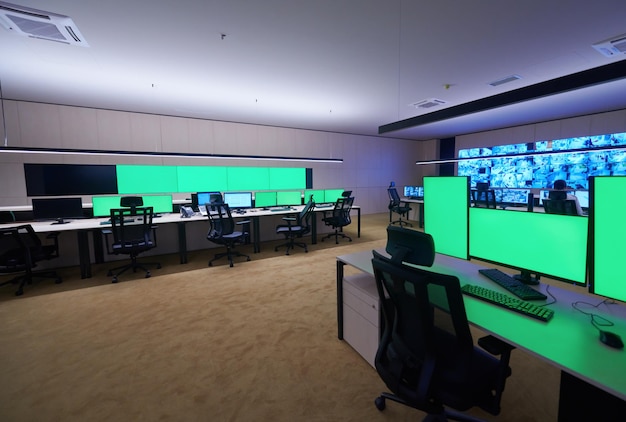Empty interior of big modern security system control room with blank green screens, workstation with multiple displays, monitoring room with at security data center  Empty office, desk, and chairs at