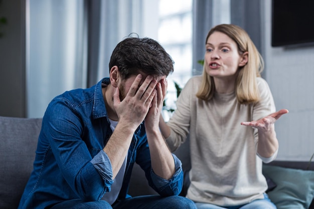 family conflict quarrel misunderstanding the woman shouts at her husband in despair crying requires explanation the man listens covering his face with his hands sitting on the couch at home