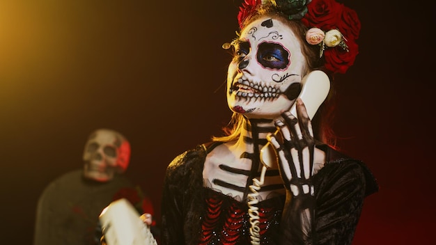 goddess of death holding landline phone in studio, talking on office telephone with cord and wearing skull make up. woman in santa muerte costume celebrating holy mexican ritual. handheld shot.