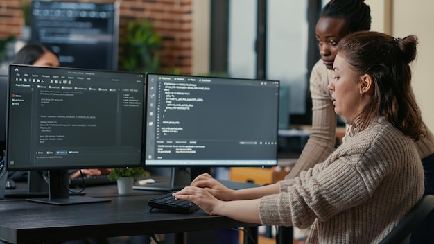 Junior programer writing algorithm under experienced colleague supervision sitting at desk with multiple screens showing programming interface. Developer writing code with the help of colleague.