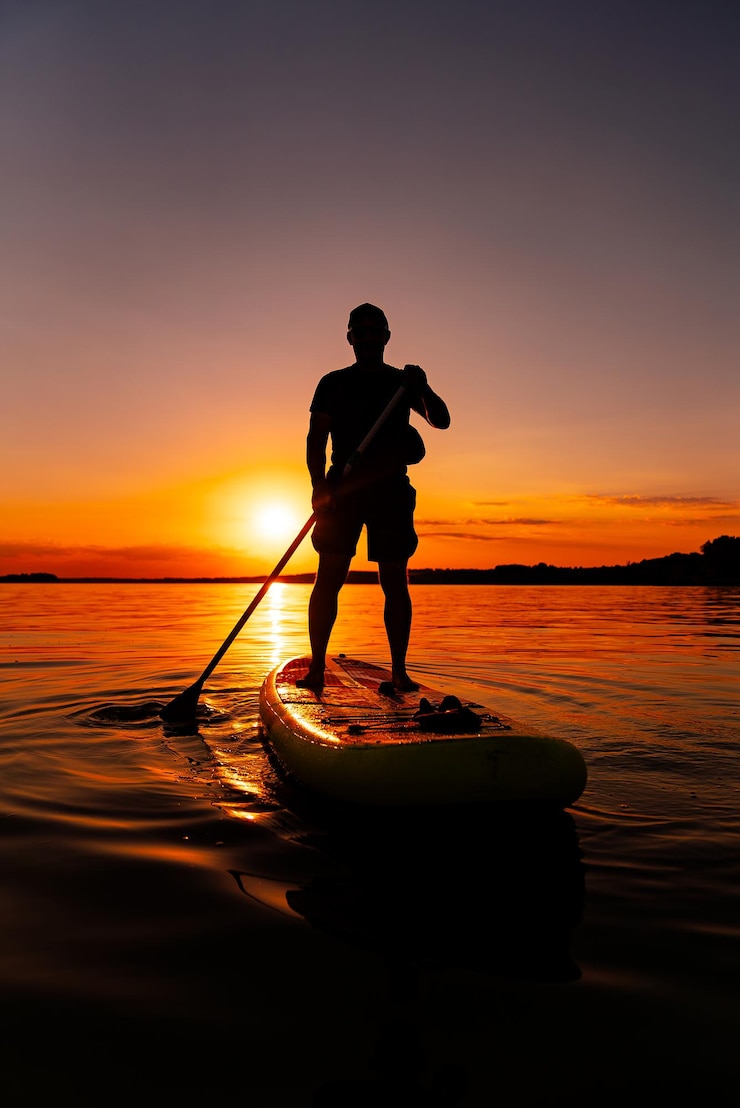 Fotografie man paddleboarding at sunset a silhouette of a man standing on a paddleboard in calm water at sunset holding a paddle