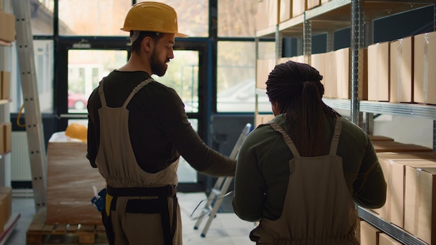 Meticulous employee walking through warehouse aisles scanning cardboard boxes on shelves under team leader guidance using digital devices to update info before sending packages for shipping