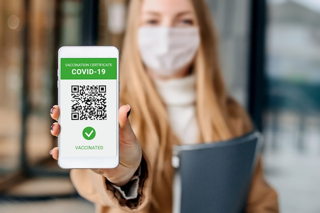 Mladá podnikatelka ukazuje obrazovku mobilního telefonu s aplikací o očkování proti koronaviru, covid-19 na pozadí budovy