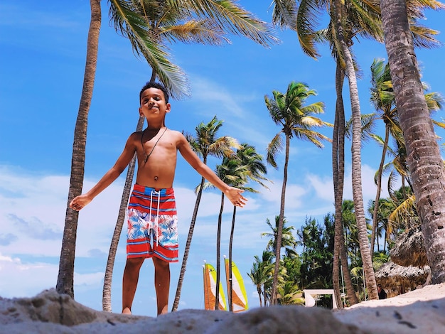 portrait of shirtless boy standing at beach against palm trees