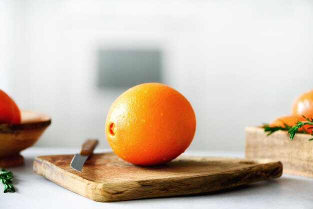 Pozadí s citrusovými plody. Čerstvé ovoce, rozmarýn. Velký pomeranč s kapkami vody, nůž, dřevěné prkénko na designové bílé kuchyni. Prostor pro text